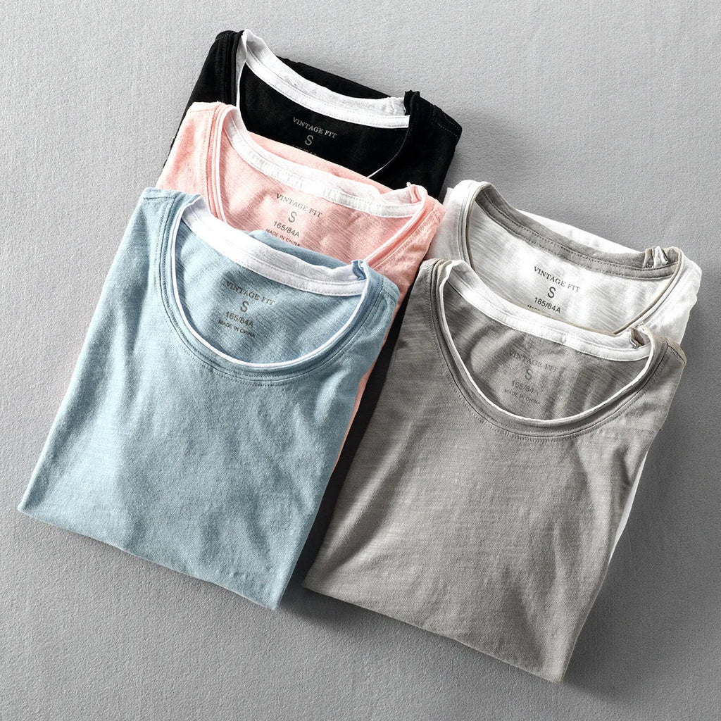Four folded t-shirts in black, pink, blue, and gray on a gray background