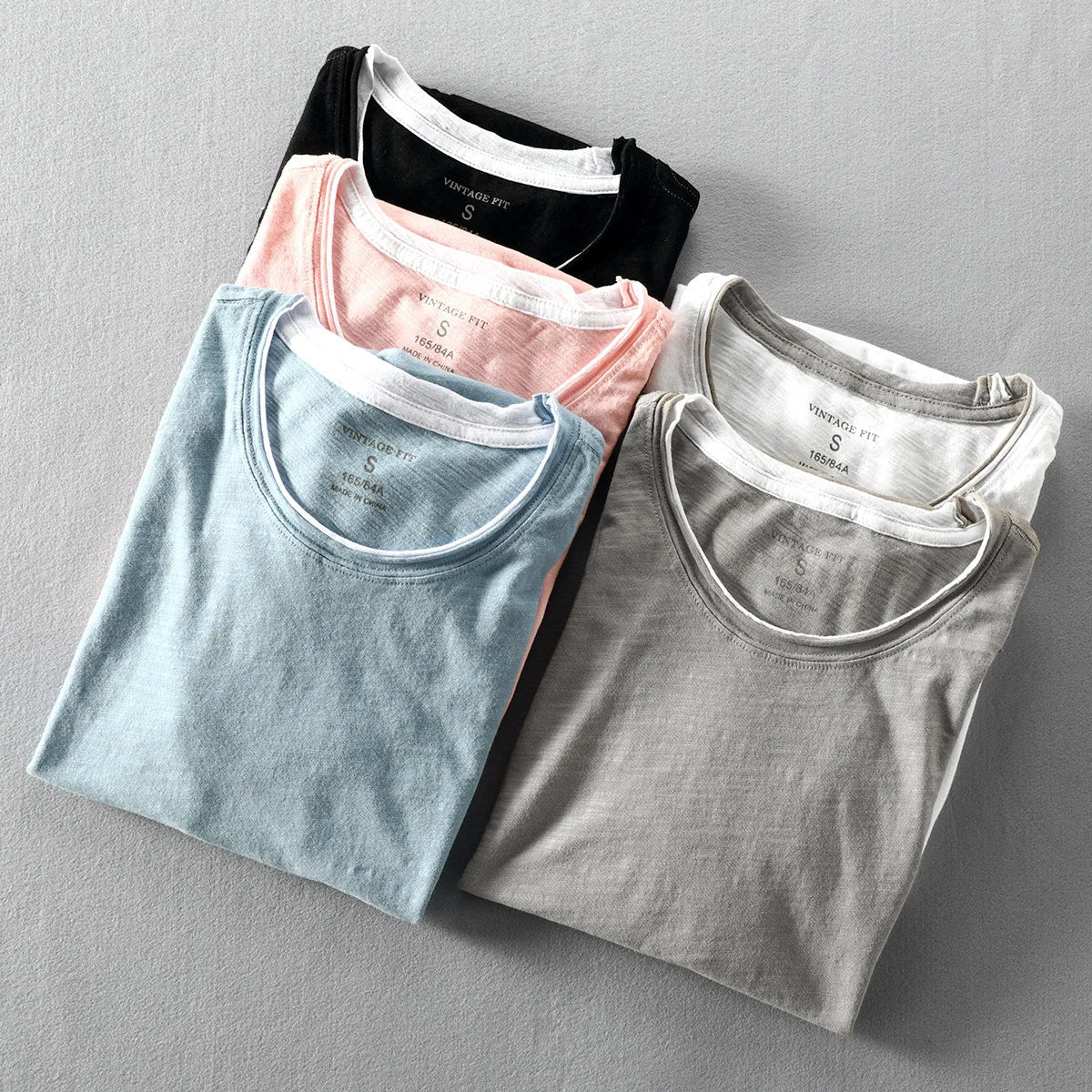 Four folded t-shirts in black, pink, blue, and gray on a gray background