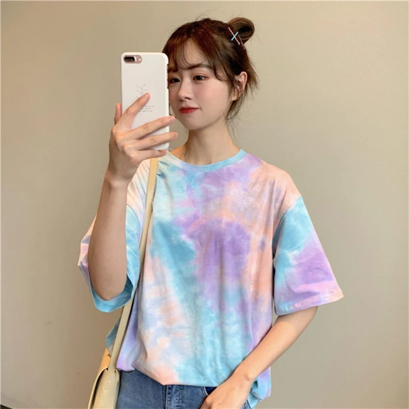 Model wearing a colorful tie dye t-shirt in pastel shades, holding a phone and smiling, showcasing a trendy summer style.