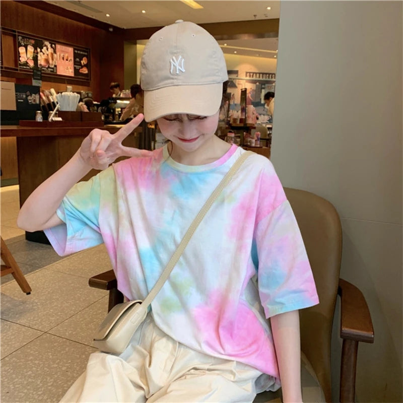 A young woman wearing a colorful tie-dye t-shirt and a beige cap, posing with a peace sign in a cafe.