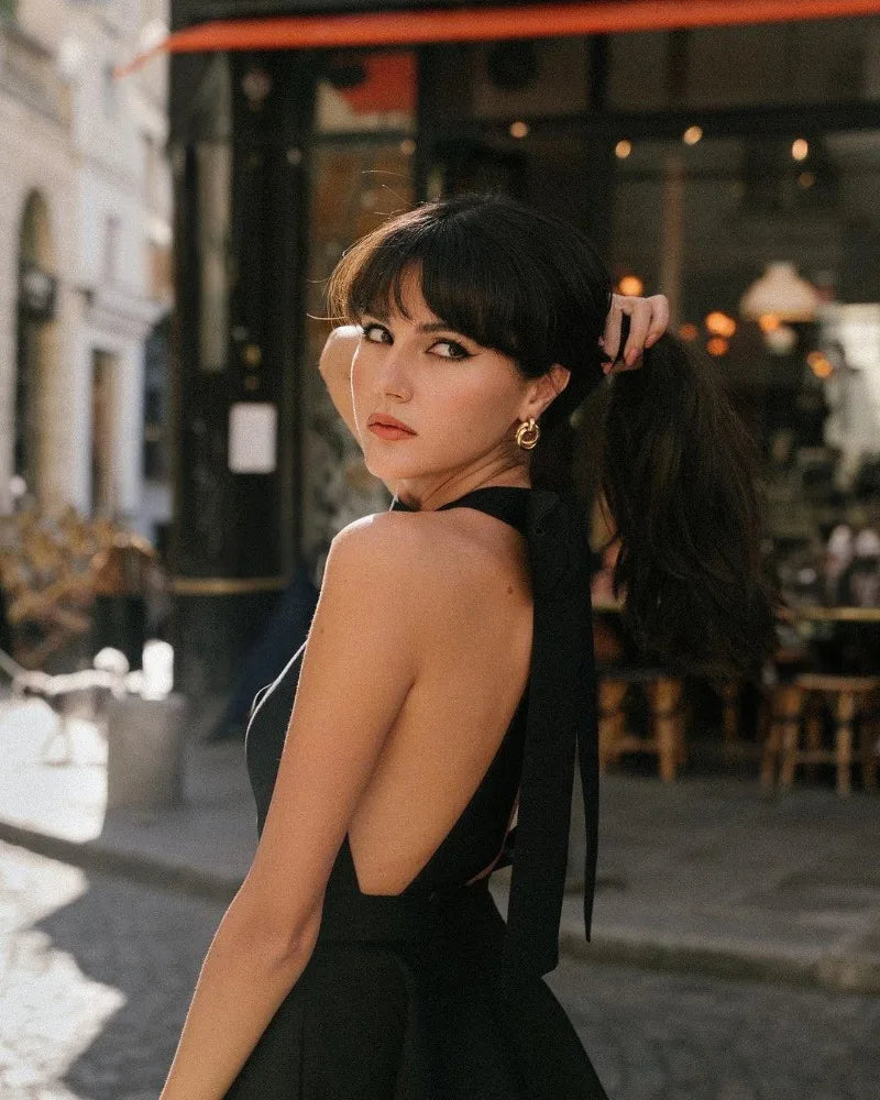 Woman in a halter dress with an open back, styled with elegant hair and earrings, posing in a sunlit outdoor café.