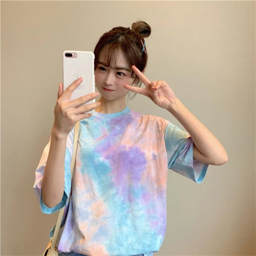 Young woman wearing a colorful tie dye t-shirt, posing for a selfie with a peace sign in front of a neutral background.