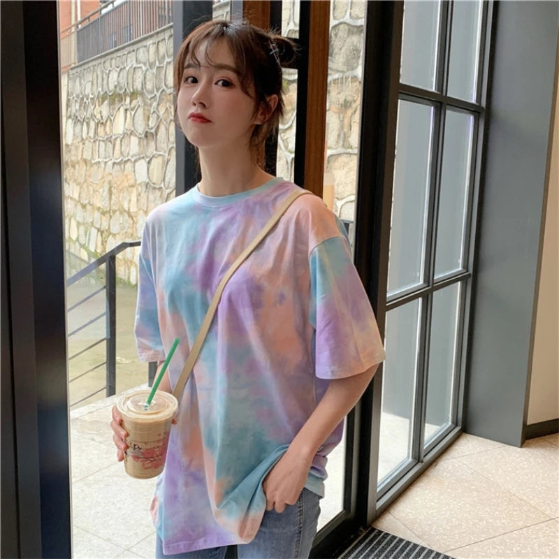 Model wearing a colorful tie dye t-shirt and holding a drink, standing near large windows.